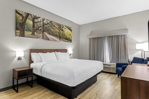 A hotel room with a bed, bedside tables, a desk, a chair, a window with curtains, and a large painting on the wall.