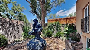 A statue of a seated figure on a sphere in a garden with a wooden fence, trees, and a building with a balcony.