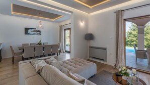 A modern living room with a large beige sectional sofa, a dining area with chairs, a flat-screen TV, and a view of an outdoor pool area. - Modern Villa Anima with swimming pool (Pazin)