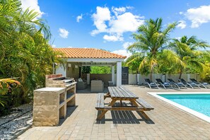 A poolside area with a wooden picnic table, lounge chairs, and a covered seating area. - Villa Noukasa, Brakkeput Abou, Lovely family home fully equipped with private pool, max 6 pers (Jan Thiel)