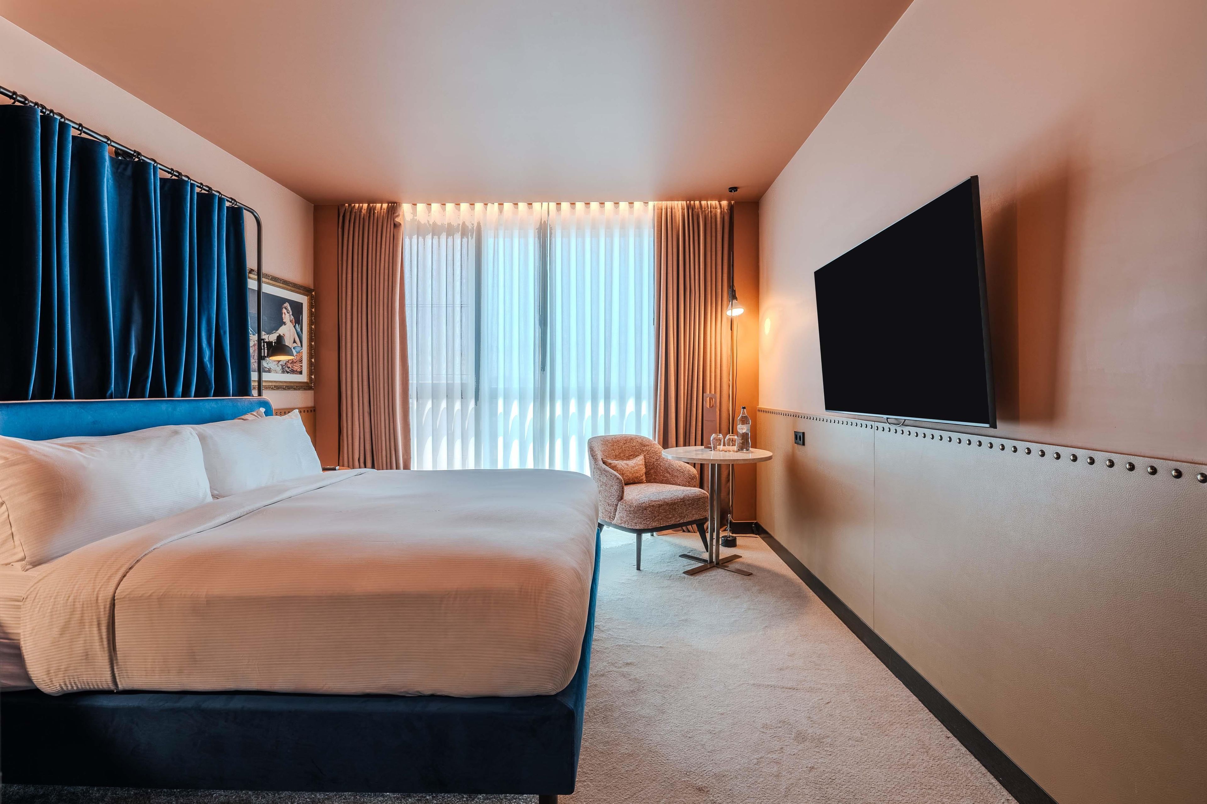 A hotel room with a bed, a chair, a small table, a TV, and curtains.