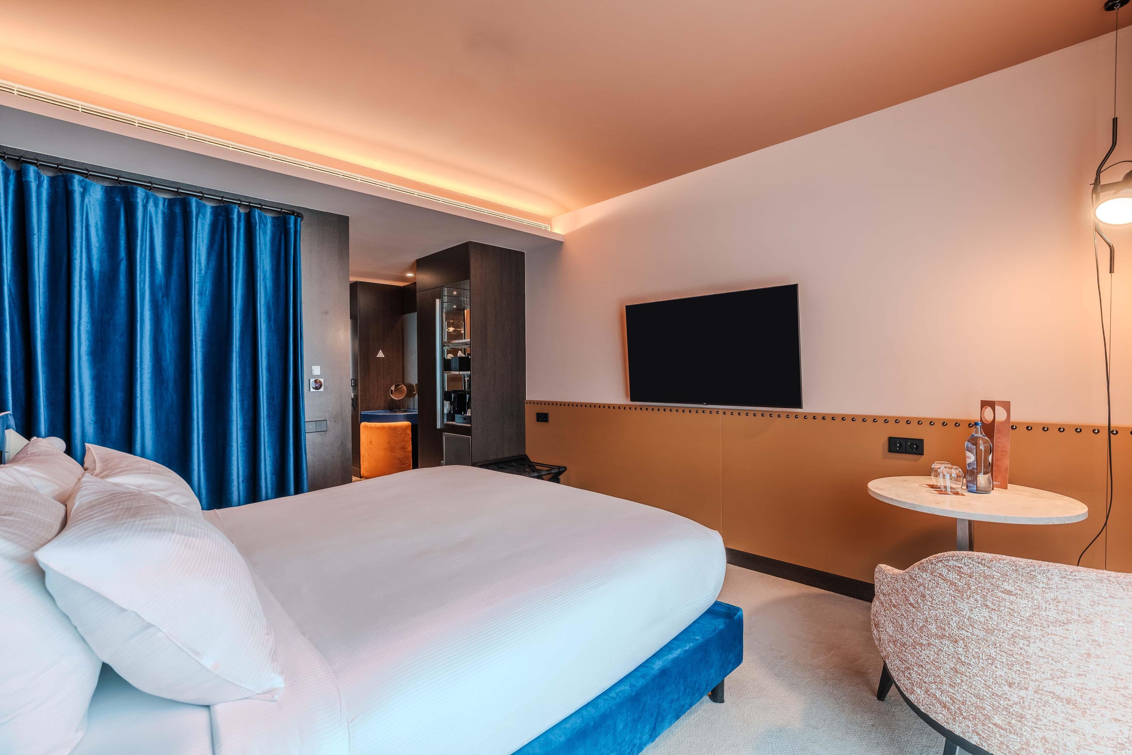A hotel room with a large bed, a small table, a TV mounted on the wall, and a bathroom visible through an open door.