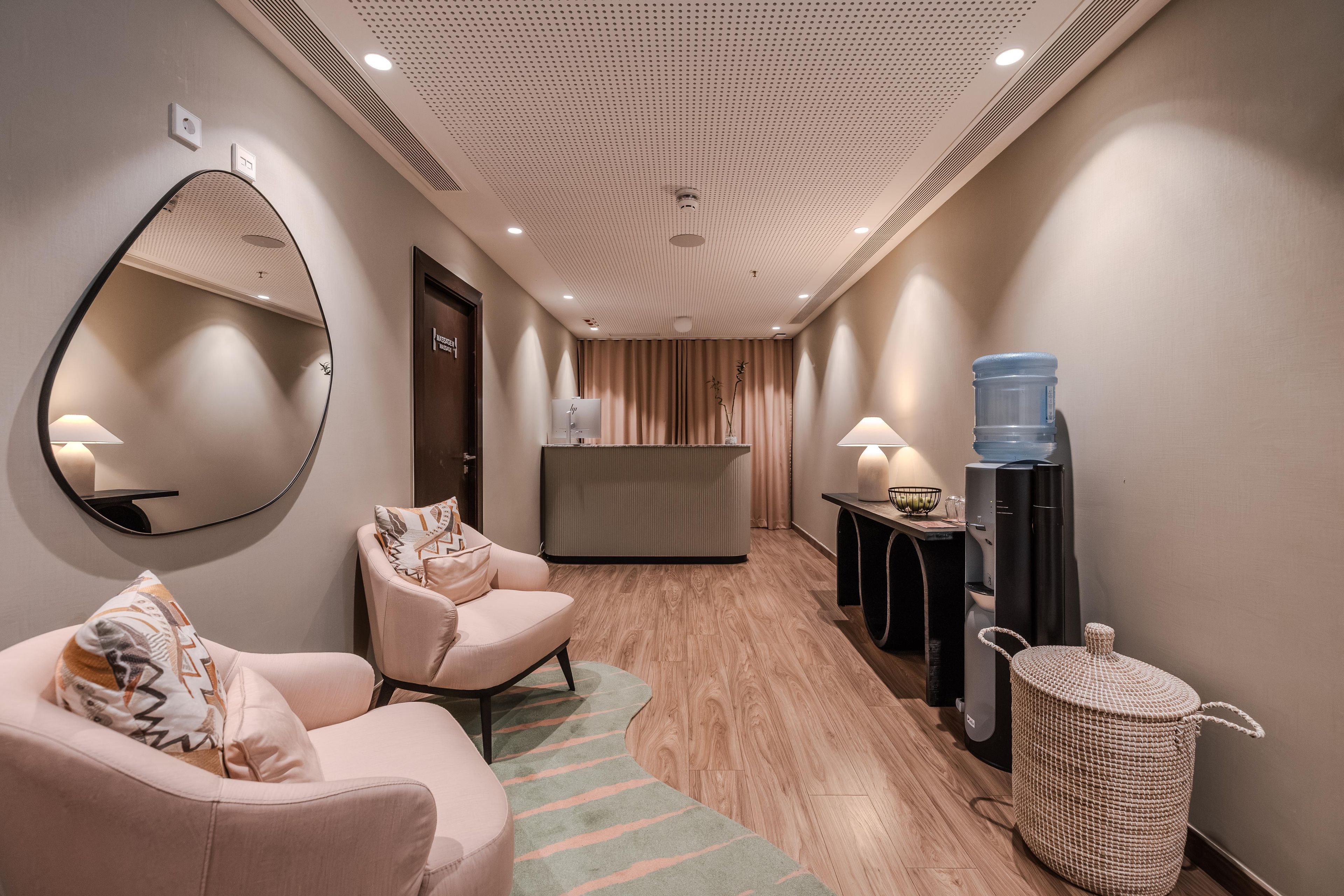 Een hotel lobby met een receptiebalie, een grote spiegel, een zithoek met een bank en stoelen, een waterkraan en een prullenbak.