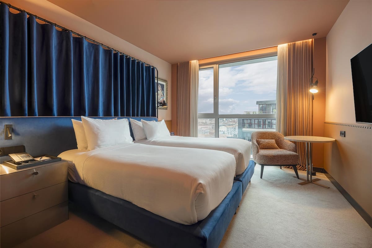 Habitación de hotel con dos camas, un escritorio, una silla, una mesita y un ventanal grande con cortinas.