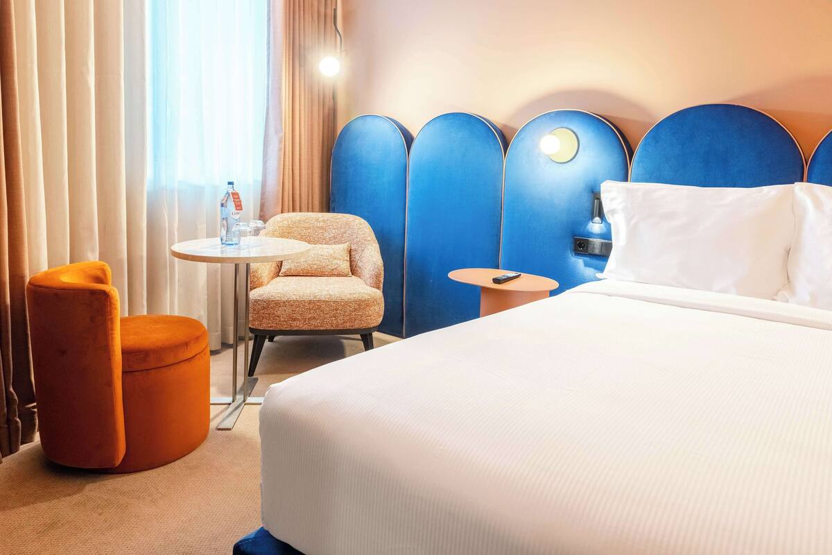 Habitación de hotel con una cama, una mesa redonda, un otoman naranja, una silla y un cabecero azul con luces.