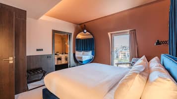 A hotel room with a large bed, a bathroom with a shower, a circular mirror, and a view of buildings through the window.