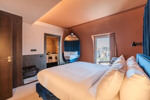 A hotel room with a large bed, a bathroom with a shower, a circular mirror, and a view of buildings through the window.