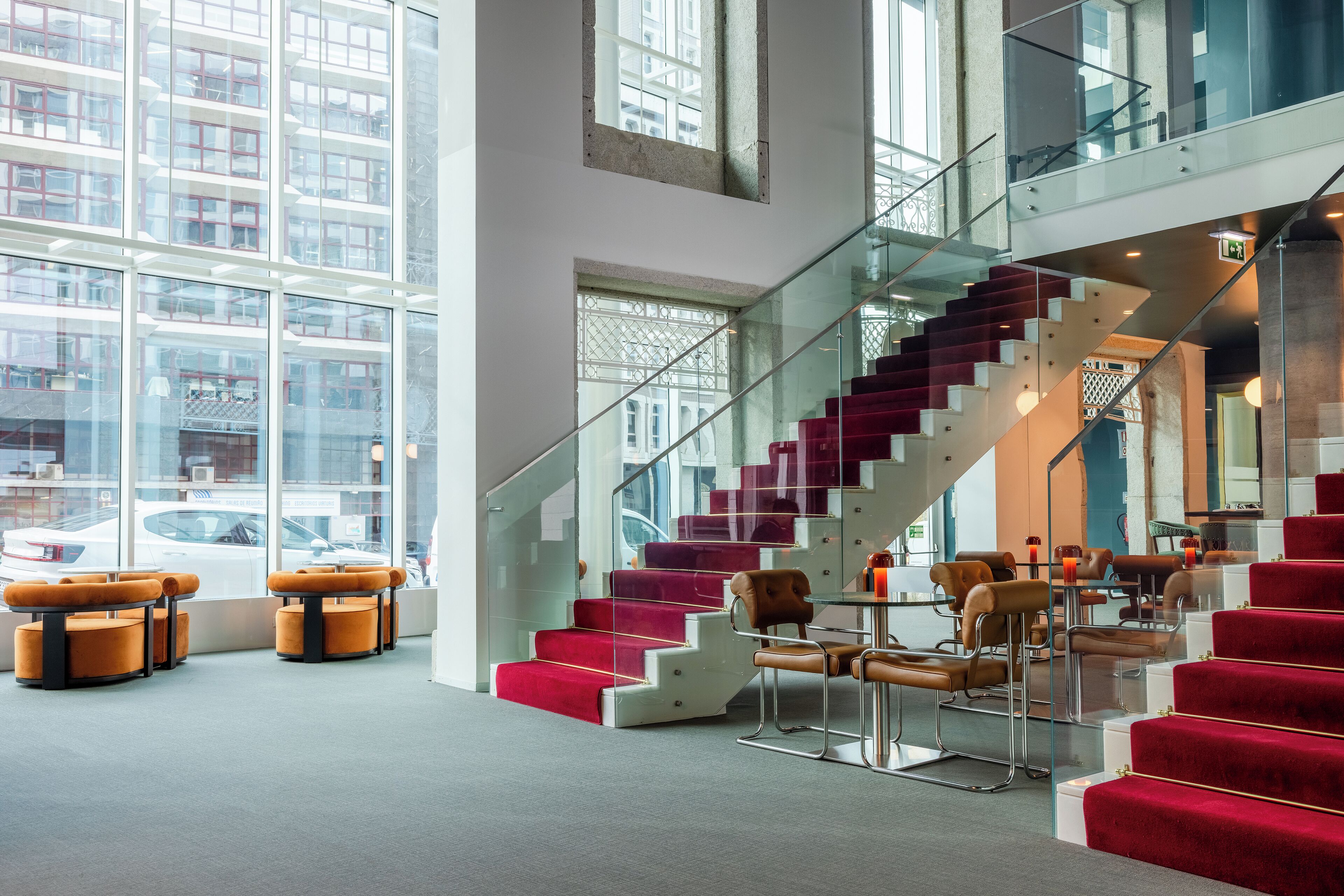 Un interior moderno con una escalera de cristal, peldaños cubiertos de alfombra roja y zonas de estar con sillas anaranjadas y marrones.