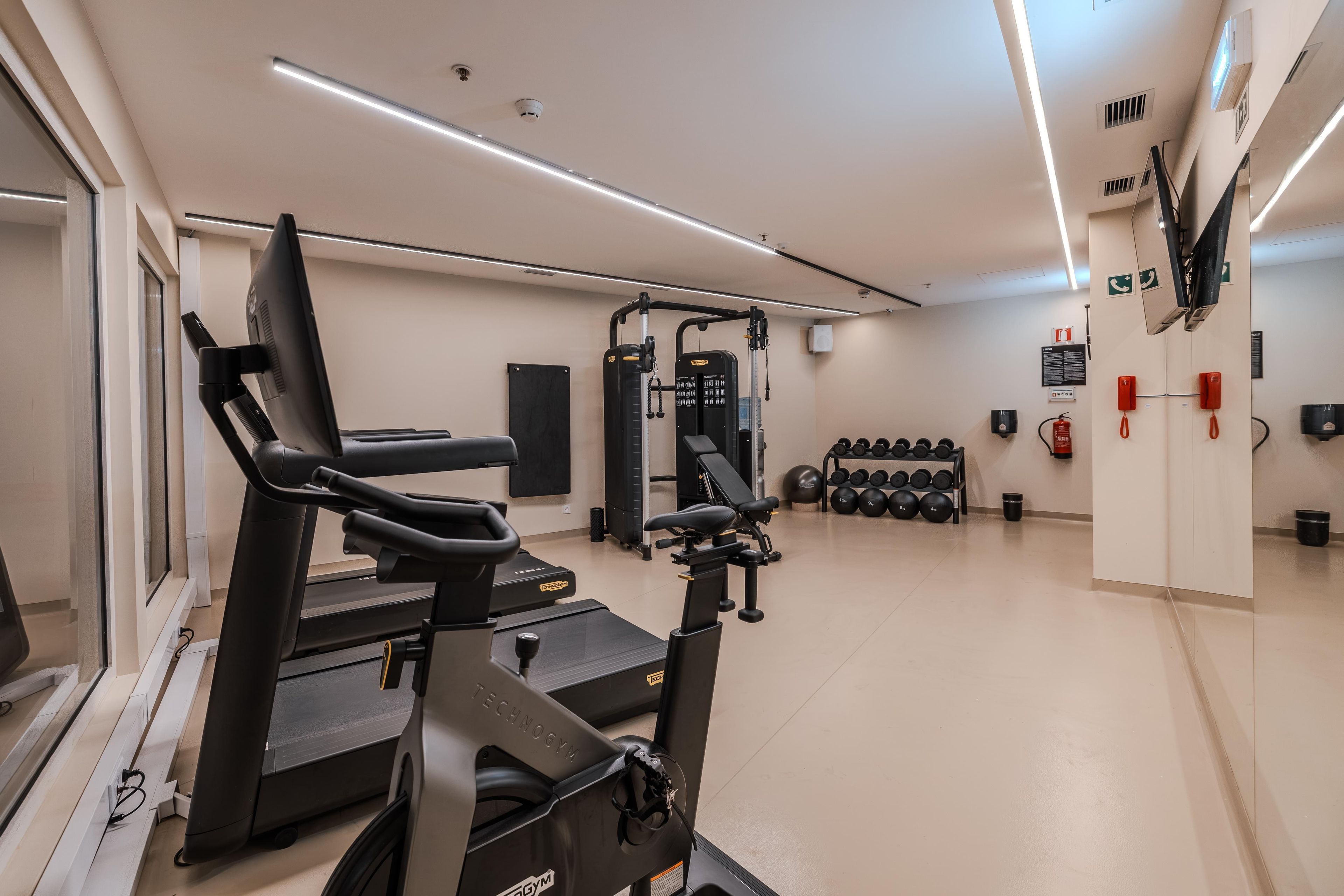Un gimnasio con equipo cardiovascular, estaciones de levantamiento de pesas y un estante para mancuernas en la pared.