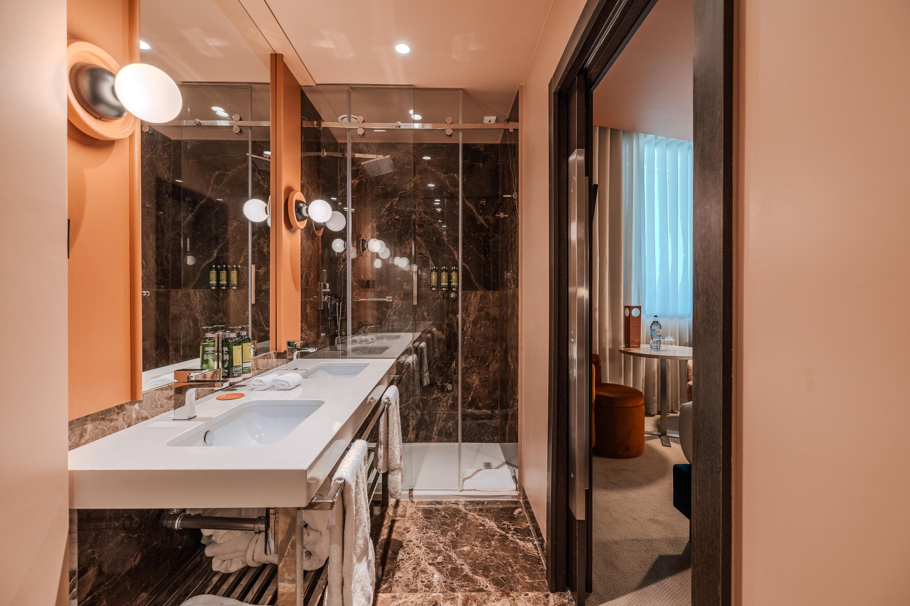 Un baño moderno con ducha envuelta en vidrio, tocador con doble lavabo y piso de mármol.