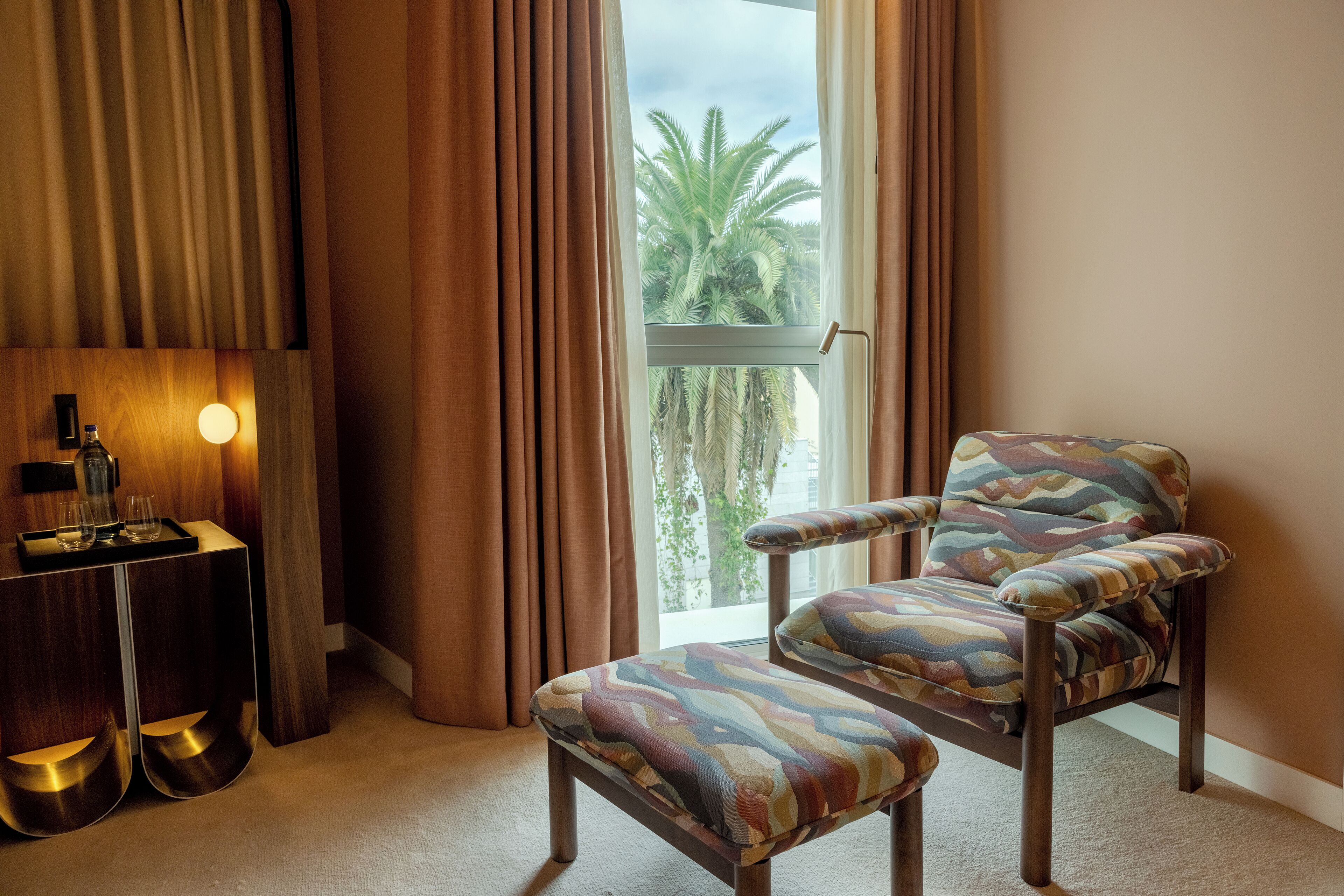 Una habitación con vista a palmeras, una silla, una mesa con bandeja y una lámpara.