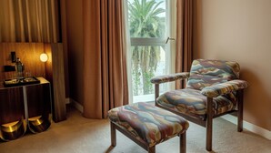 A room with a view of palm trees, a chair, a table with a tray, and a lamp.