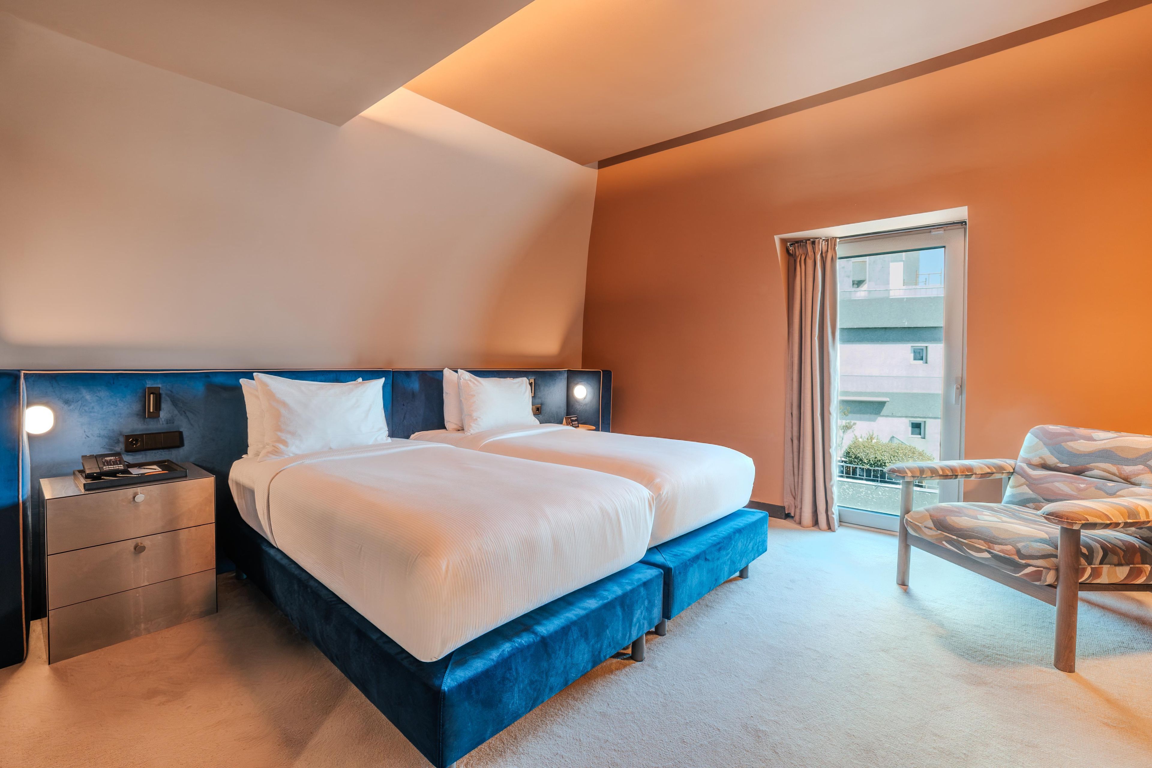 A hotel room with a large bed, a nightstand, a chair, and a window with a view of buildings.