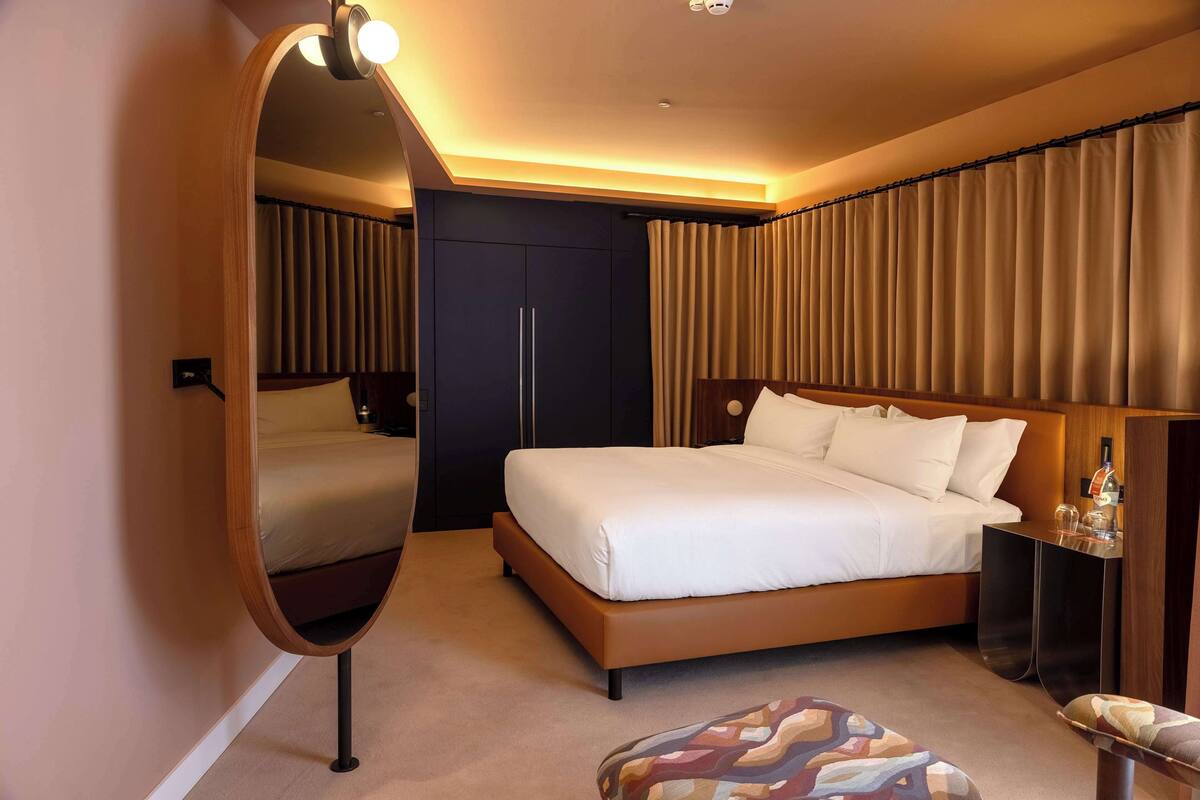 Habitación de hotel moderna con una cama grande, una mesita de noche con lámpara, un espejo y vistas a las cortinas.