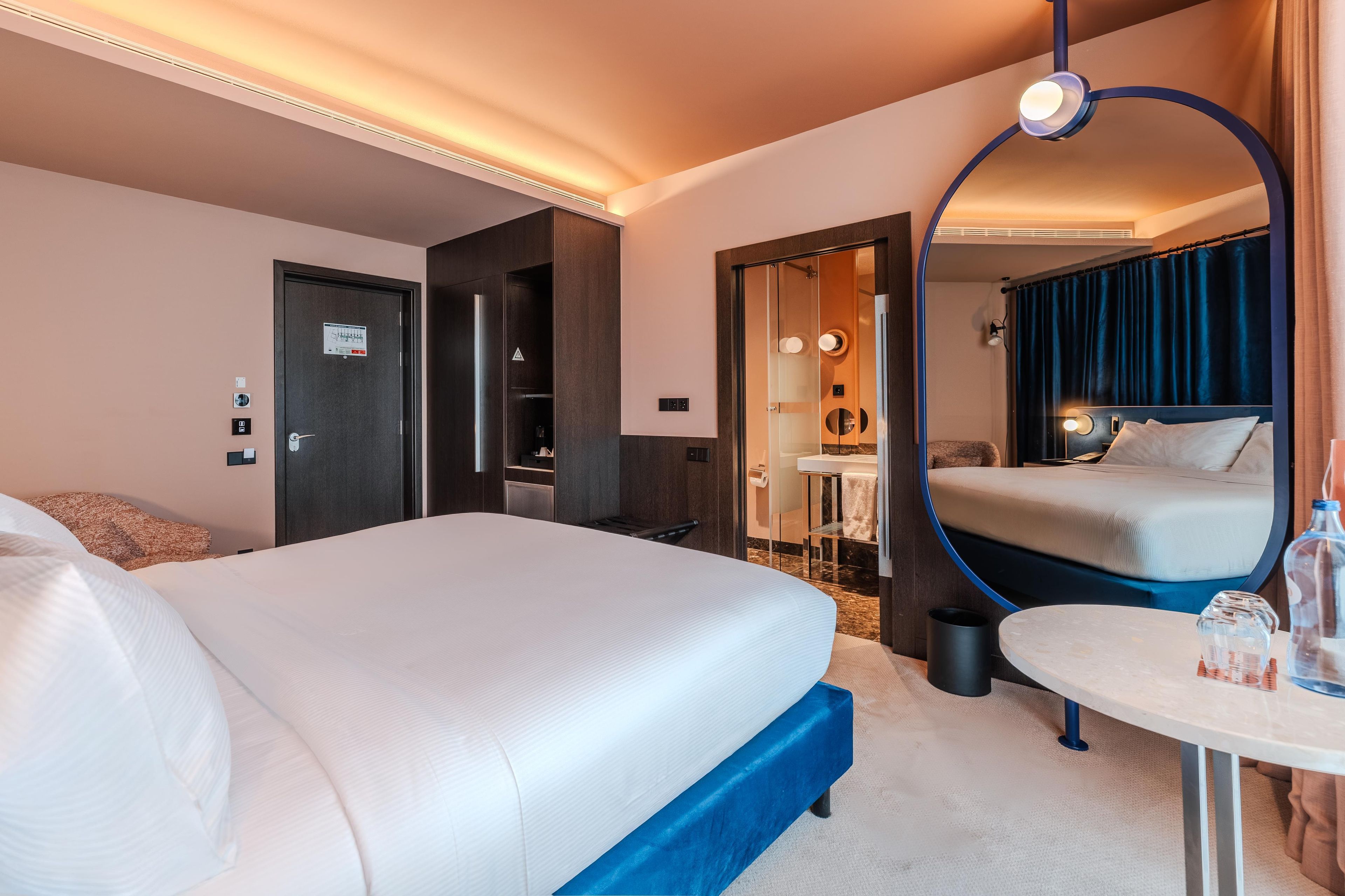 Een hotelkamer met een groot bed, een klein tafeltje, een spiegel en een badkamer die door een open deur zichtbaar is.