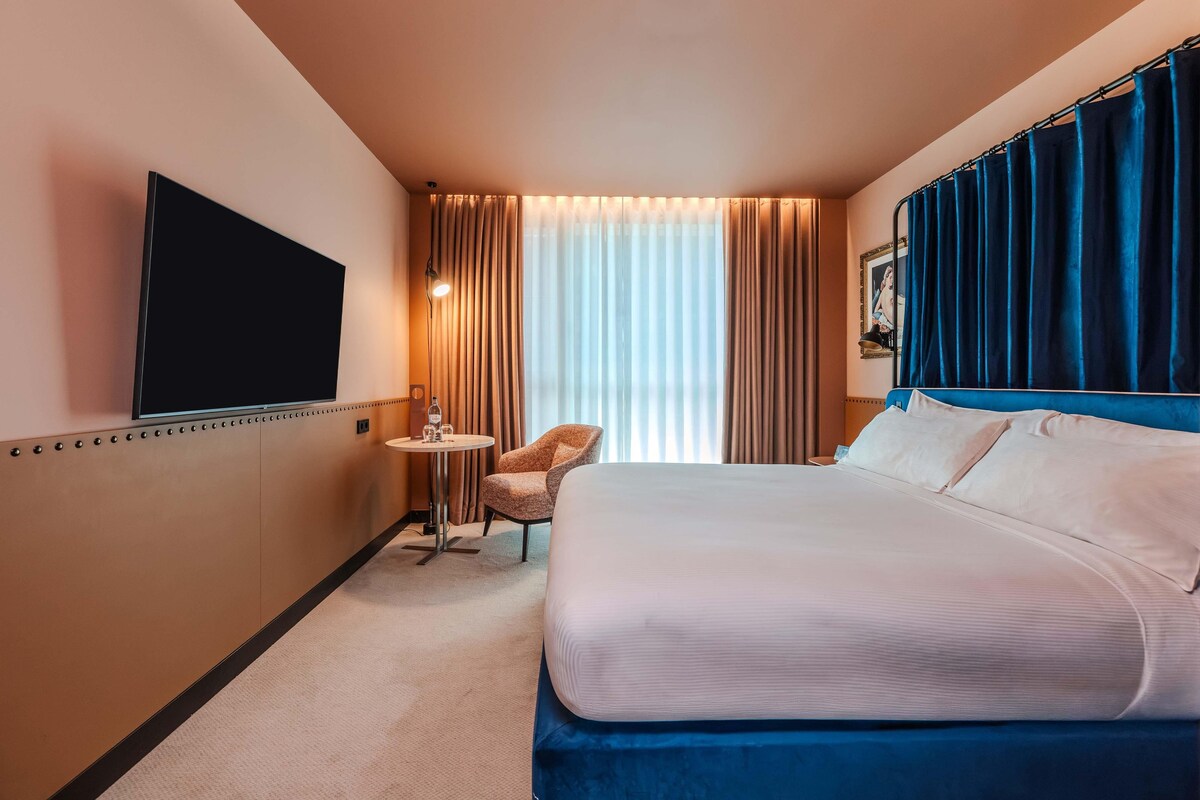 Habitación de hotel con cama, una silla, televisión, mesita y ventana con cortinas.