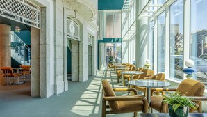 Un espacio interior moderno con una hilera larga de mesas redondas y sillas de mimbre, ventanas amplias que ofrecen vistas de la calle, y una escalera que conduce a un nivel superior.