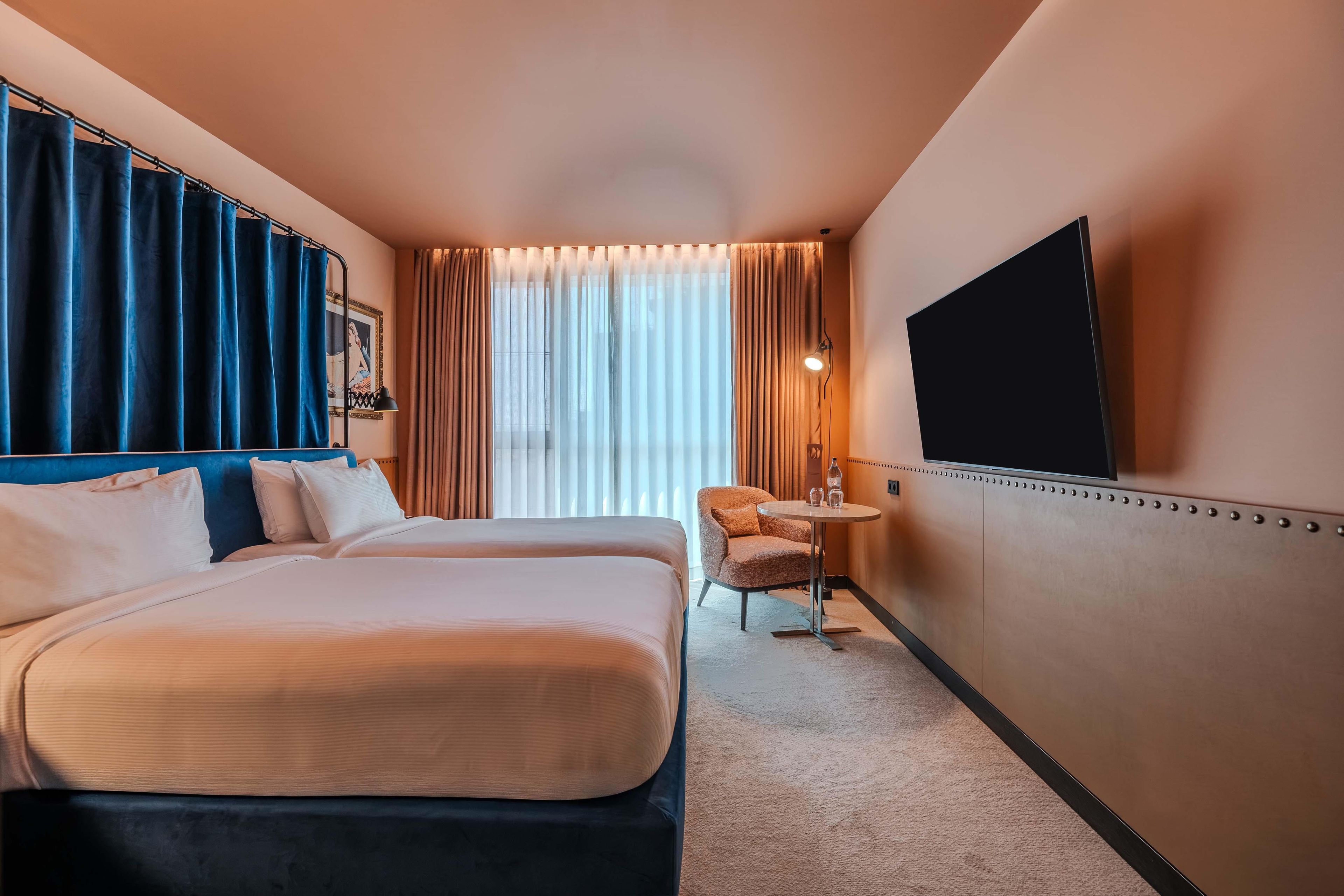 A hotel room with a bed, a chair, a small table, a TV, and a window with curtains.