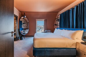Habitación de hotel con una cama grande, un espejo redondo, vistas de la ciudad y una ventana con cortinas.