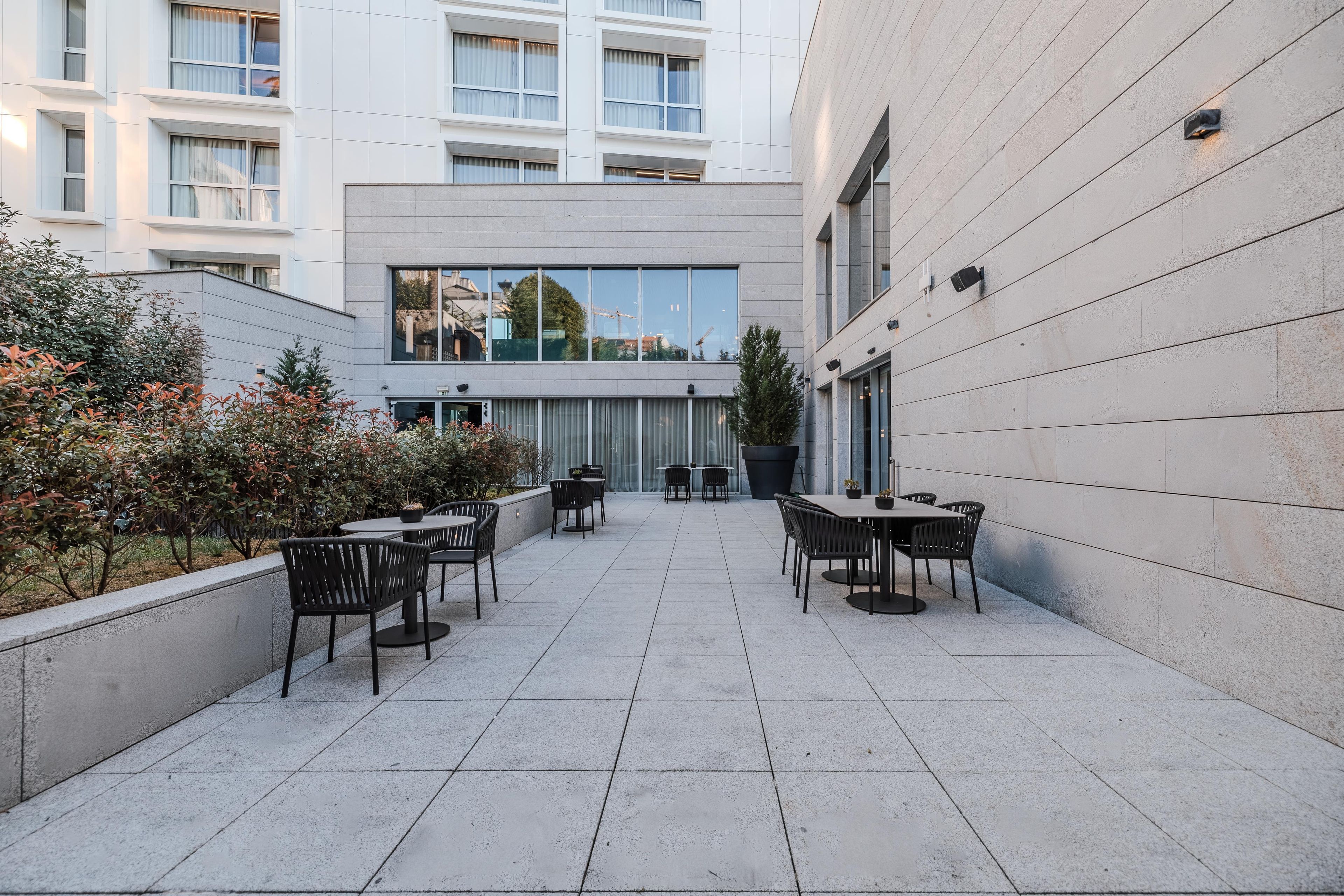 Un patio al aire libre con mesas y sillas, un edificio con grandes ventanales y vistas del entorno.