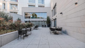 Un patio al aire libre con mesas y sillas, un edificio con grandes ventanales y vistas del entorno.