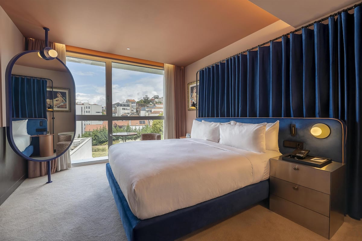 Habitación de hotel con una cama grande, una mesita de noche con lámpara, un espejo redondo y vistas a la ciudad.