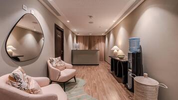 Un vestíbulo de hotel con una mesa de recepción, un espejo grande, zona de estar con sofá y sillones, dispensador de agua y un cubo de basura.
