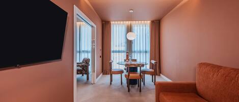 A hotel room with a flat-screen TV, a round dining table with chairs, a sofa, and a large window with curtains.