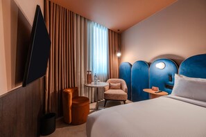 Une chambre d’hôtel avec un lit, une chaise, une petite table, un téléviseur fixé au mur et une fenêtre avec des rideaux.