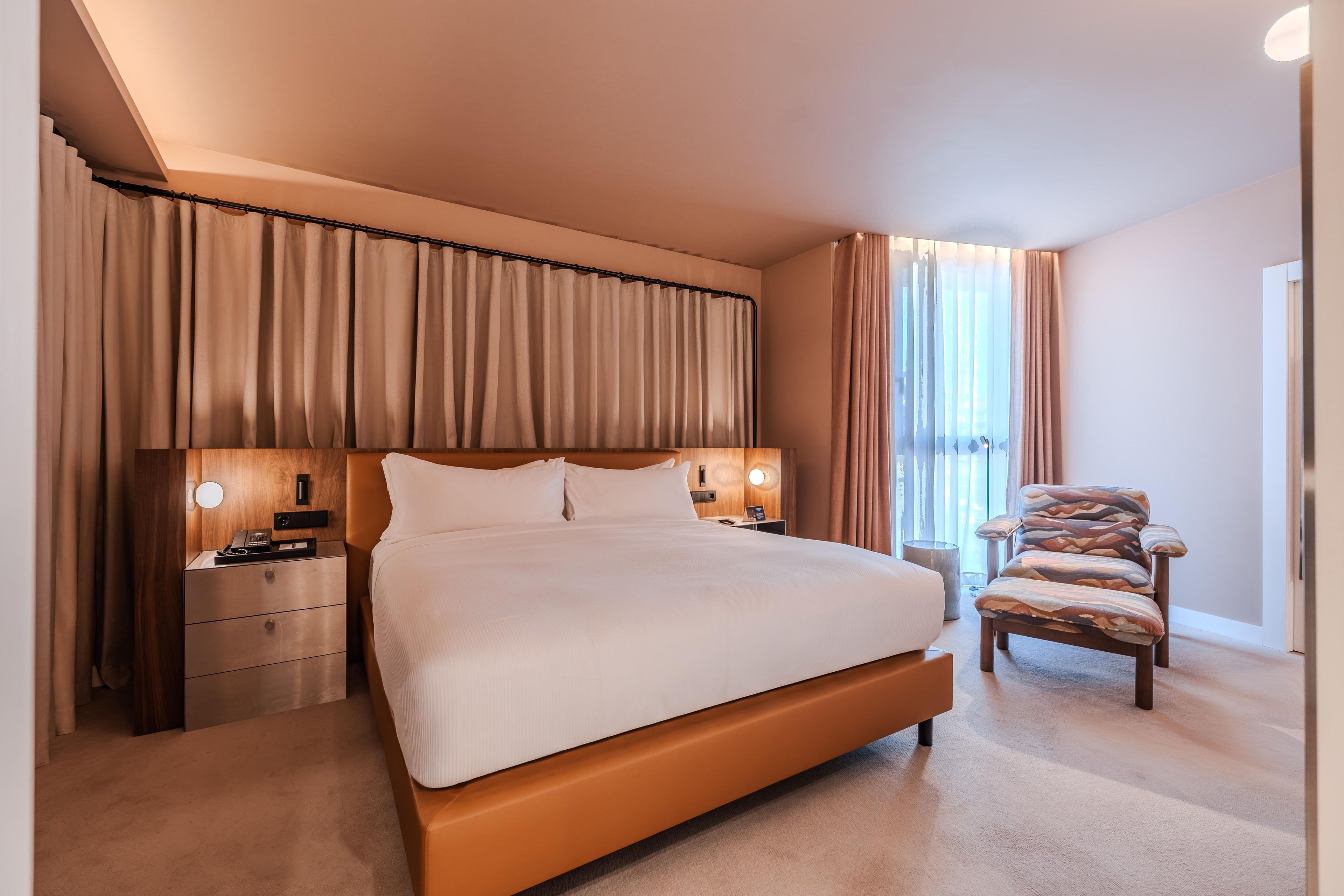 A hotel room with a large bed, bedside tables with lamps, a chair, and curtains.