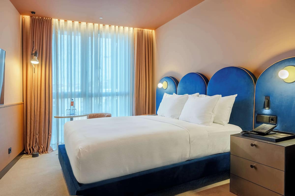 Habitación de hotel con una cama grande, una mesita de noche con lámpara y vistas a la ciudad por la ventana.