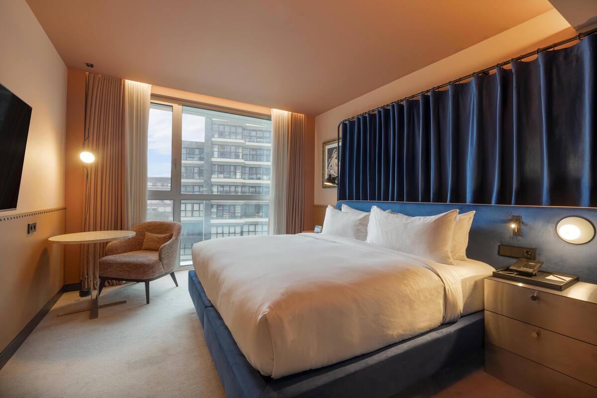 Una habitación de hotel con cama, mesita de noche, silla y un gran ventanal con cortinas.