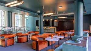 Un área de estar moderna con asientos naranja y azul, un bar y ventanas amplias.