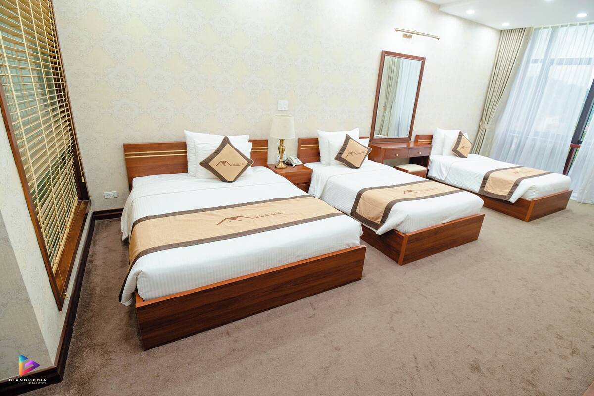 A hotel room with three beds, a wooden headboard, a bedside table with a lamp, and a large mirror on the wall.