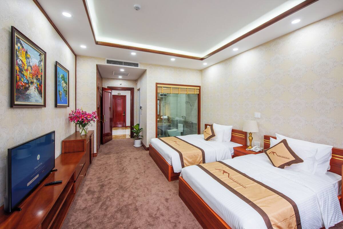 A hotel room with two beds, a TV, a bathroom with a glass shower, and a hallway leading to another room.