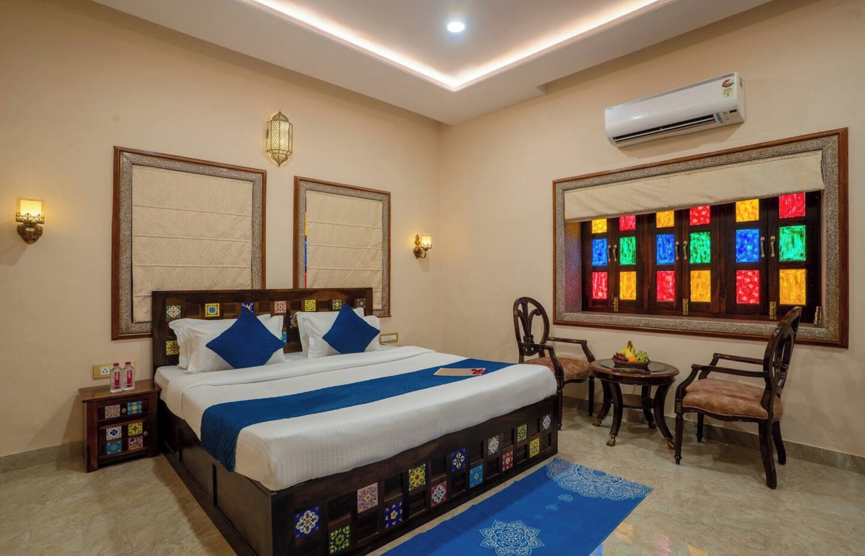 Une chambre d’hôtel avec un grand lit, deux climatiseurs fixés au mur, une fenêtre ornée de vitraux colorés, une petite table avec des fruits et un tapis bleu.