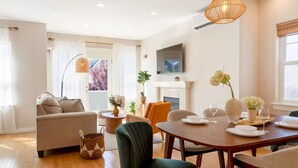 A modern living room with a sofa, dining table, and chairs. There is a fireplace, a TV mounted on the wall, and a large window with curtains. - Spacious 3B/2.5B Home in the Heart of Capitol Hill (Seattle)
