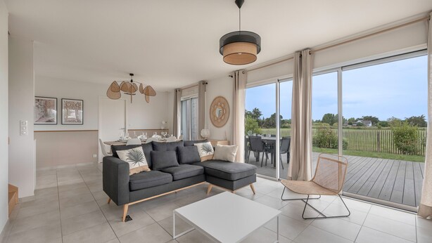 A modern living room with a grey sectional sofa, a white coffee table, and a wicker chair. There is a large window offering a view of a garden and a house. - 650m from the sea, modern house for 7 (Piriac-sur-Mer)