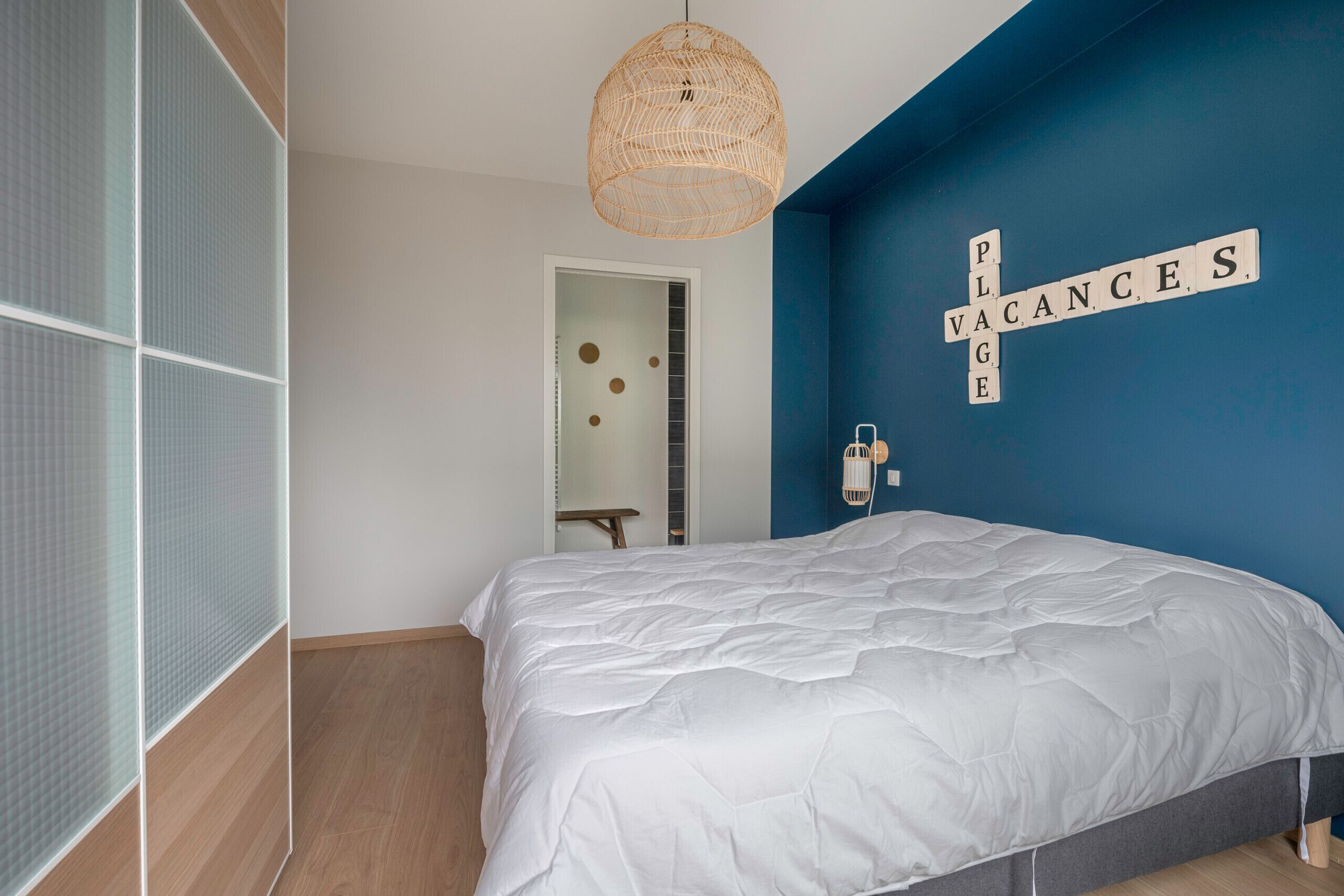 A bedroom with a large bed, a blue accent wall, and a decorative sign spelling "VACANCES".