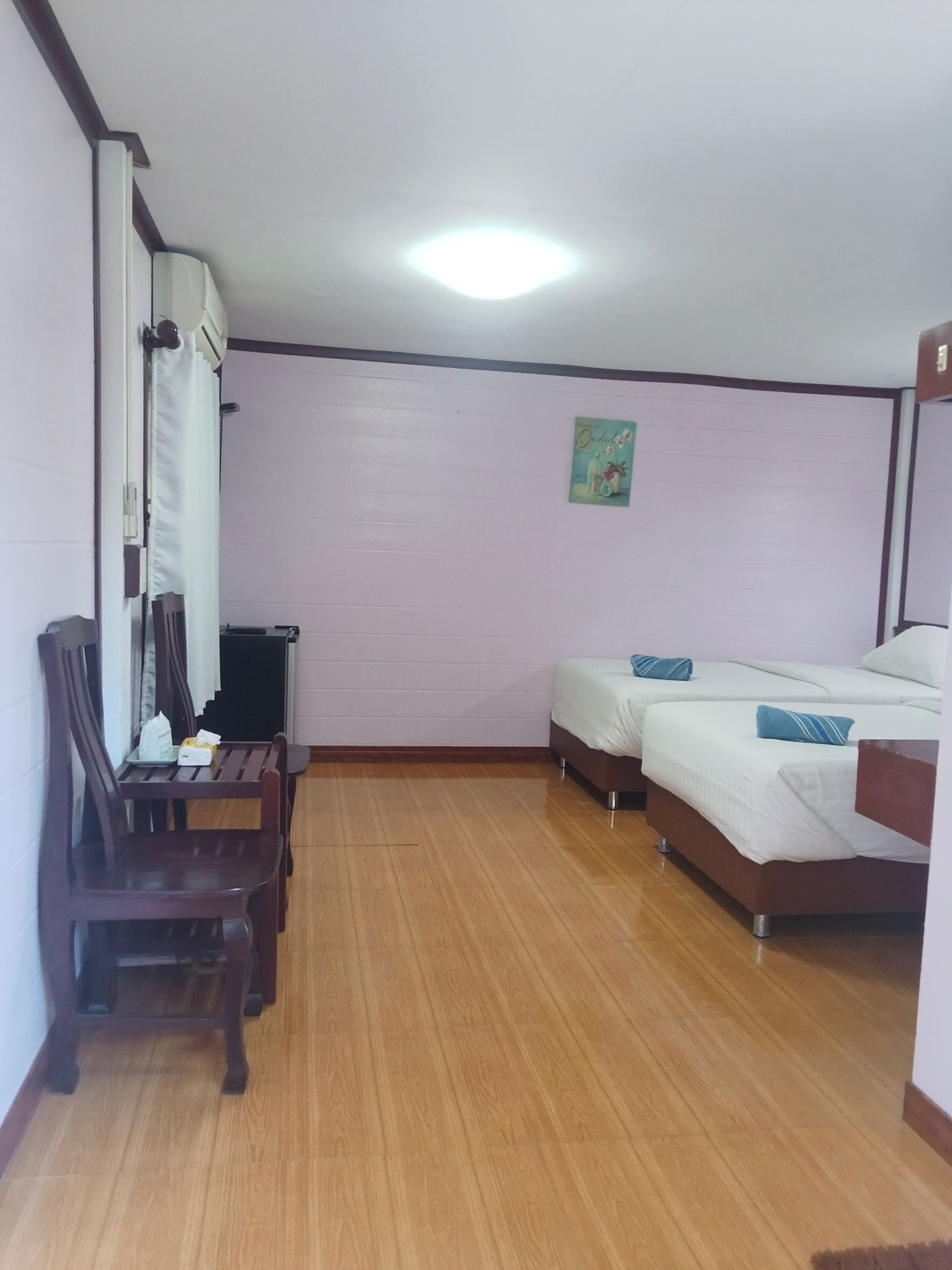 A hotel room with two single beds, a wooden desk with a chair, a small table, and a wall-mounted air conditioner.