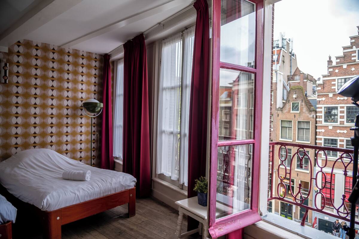 A room with a bed, a window with curtains, a small table with a plant, and a patterned wall.