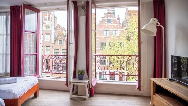 Una habitación con una cama, una mesita con una planta, un escritorio con una lámpara y ventanas amplias con cortinas rojas.