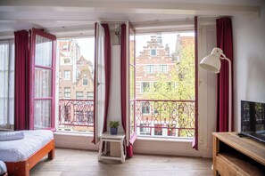 A room with a bed, a small table with a plant, a desk with a lamp, and large windows with red curtains. - Hotel & bar Royal taste Amsterdam (Amsterdam)