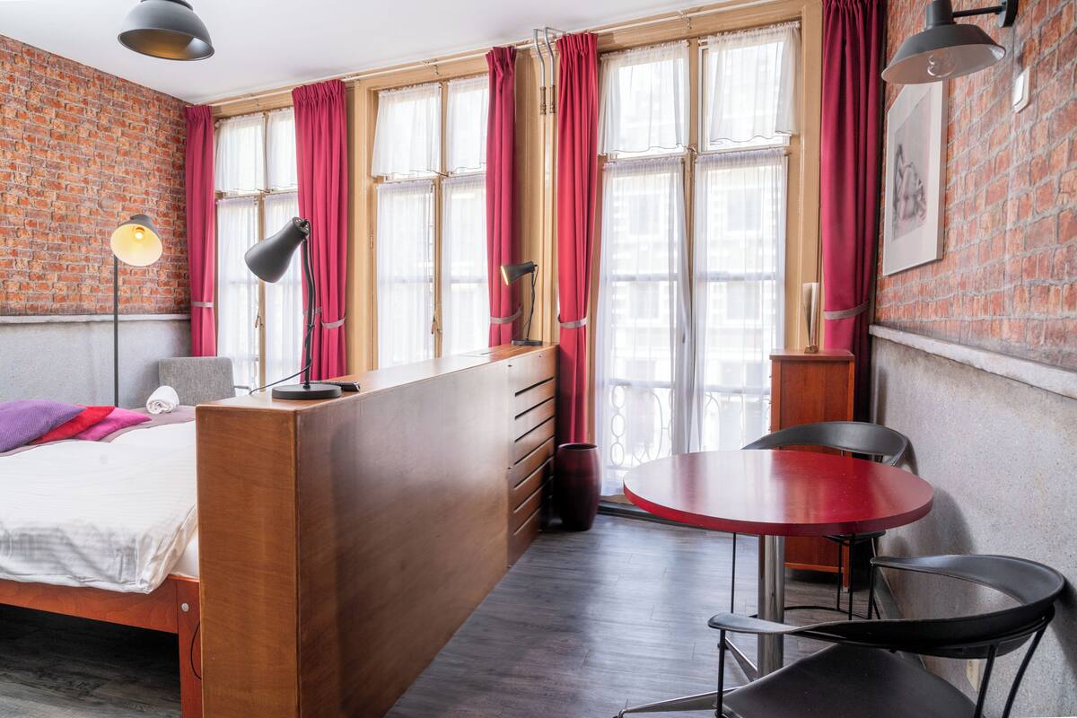 A room with a wooden bed, a desk, a round red table, and chairs. There are windows with red curtains and a brick wall.