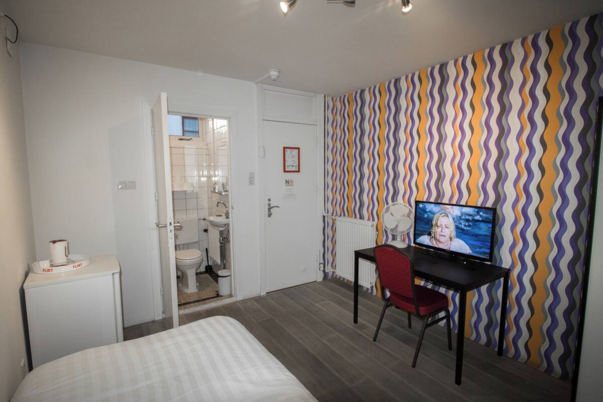 A hotel room with a bed, a desk with a chair, a television, and a bathroom visible through an open door.