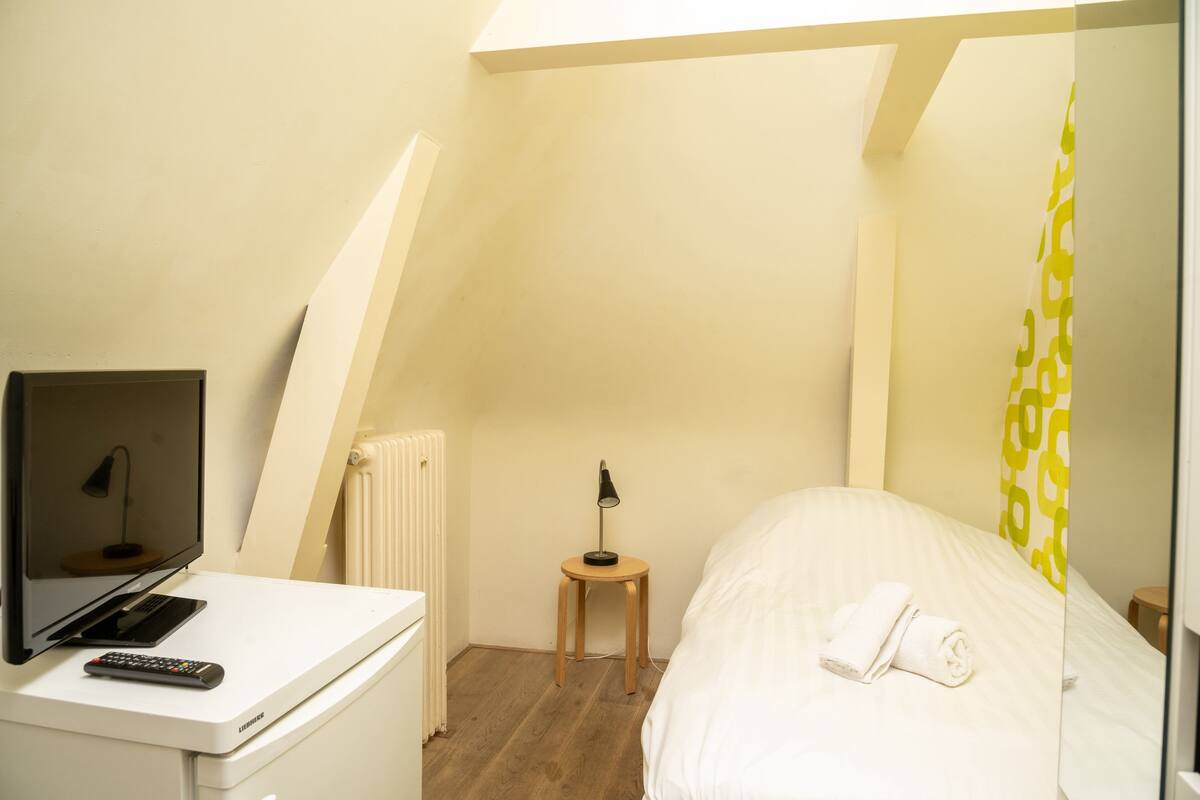 A small, well-lit room with a bed, a desk with a computer and a lamp, and a radiator.