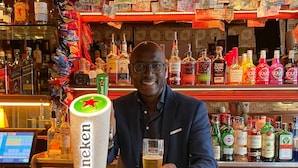 Un cantinero sosteniendo una cerveza Heineken en un bar con una variedad de botellas de licor y una caja registradora.