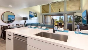 Una cocina moderna con una isla central, un área de comedor con un sofá azul y ventanas amplias que dan a vistas de árboles y un cielo despejado.