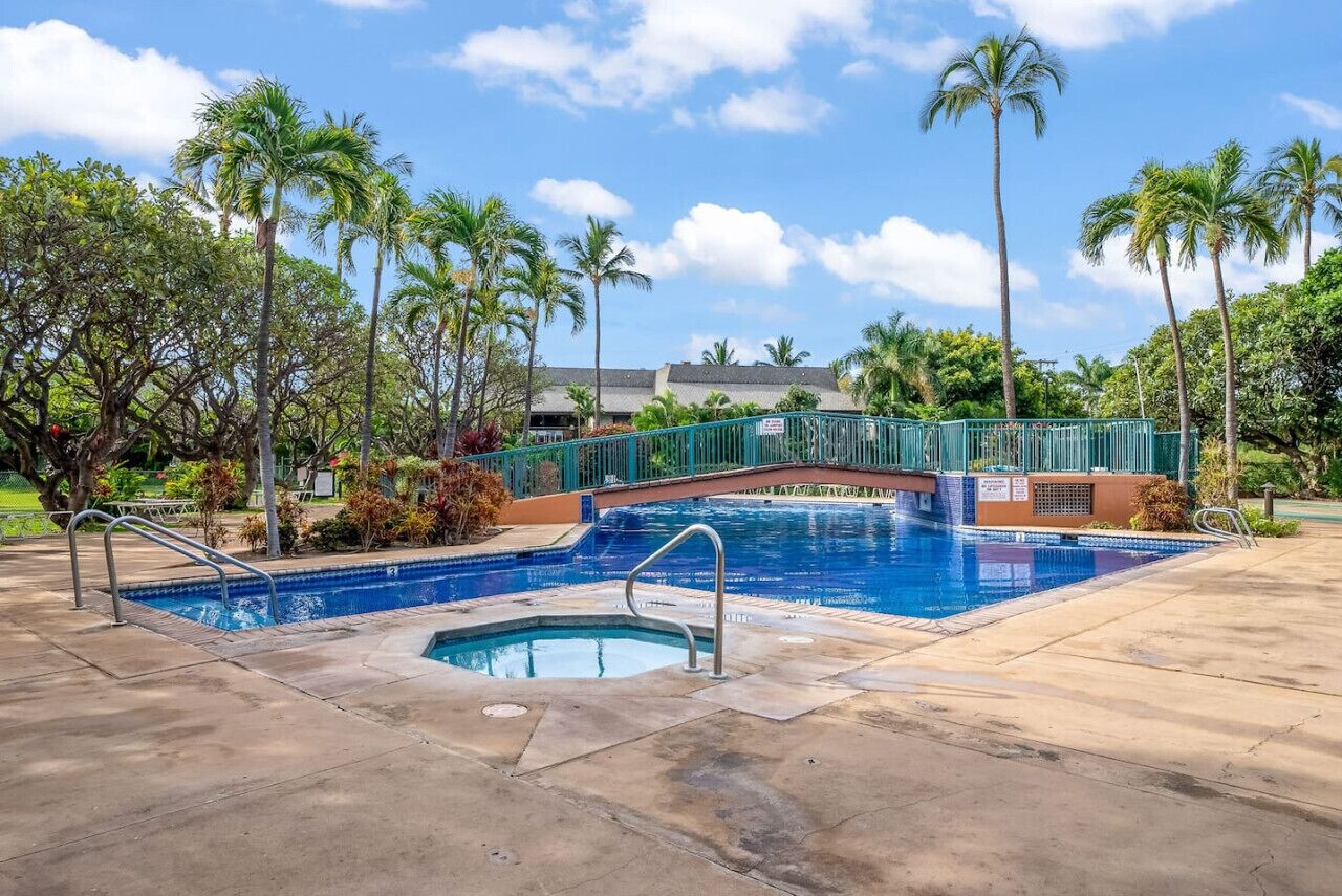 A swimming pool with a bridge, surrounded by palm trees and a building in the background.