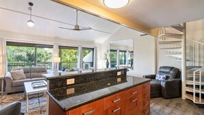 A modern kitchen with a central island, a dining area with a sofa and a coffee table, and a staircase leading to an upper level. - Steps to the Beach! 3 Bedroom! (Kihei)